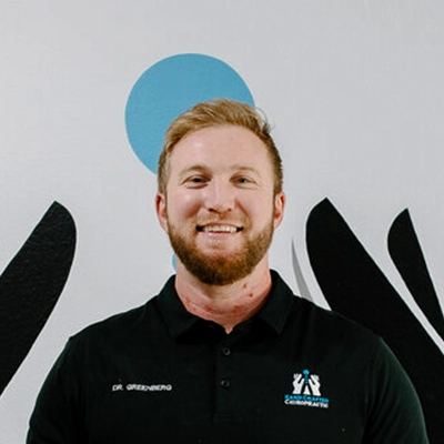Chiropractor Adam Greenberg Smiling In Front Of Logo Wall