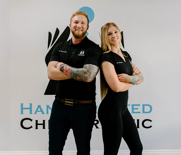 Chiropractors Adam Greenberg And Jenna Swaffer Smiling In Front Of Logo Wall